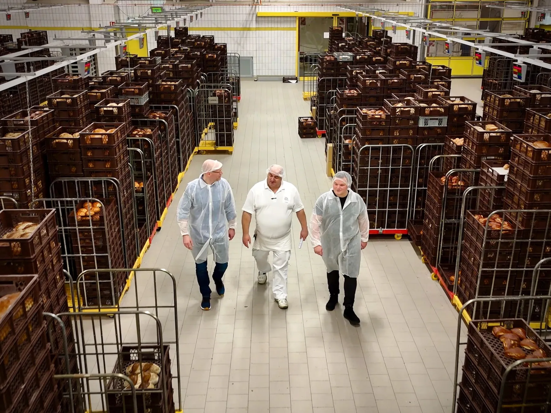 Drei Personen in Hygienekleidung gehen durch die Versandhalle einer Bäckerei zwischen gestapelten Brotkisten